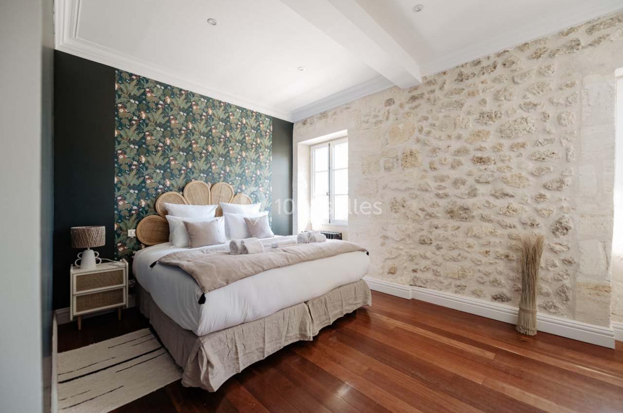 Chambre lumineuse avec lit double, tête de lit en rotin, mur en pierre apparente et papier peint à motifs végétaux.