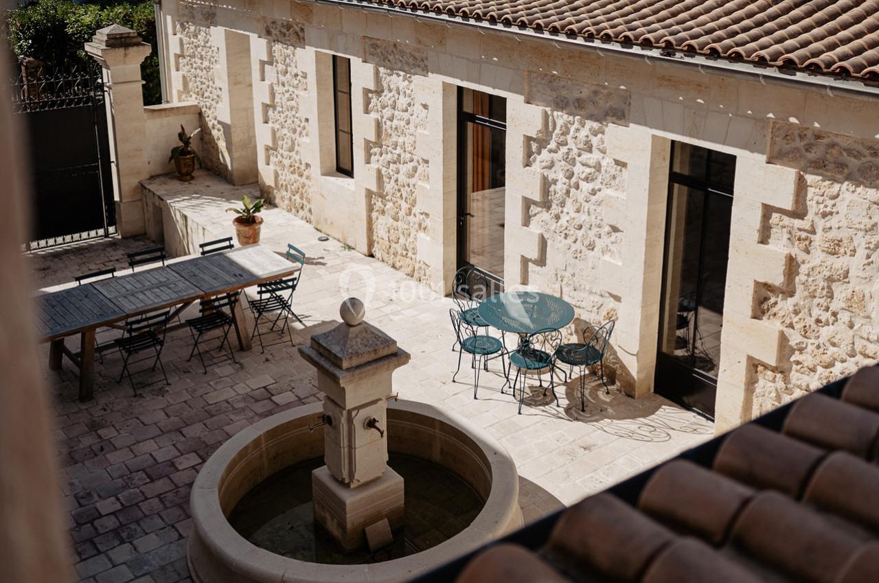 Cour intérieure en pierre avec une fontaine centrale, une grande table en bois, des chaises et des murs en pierre apparente.
