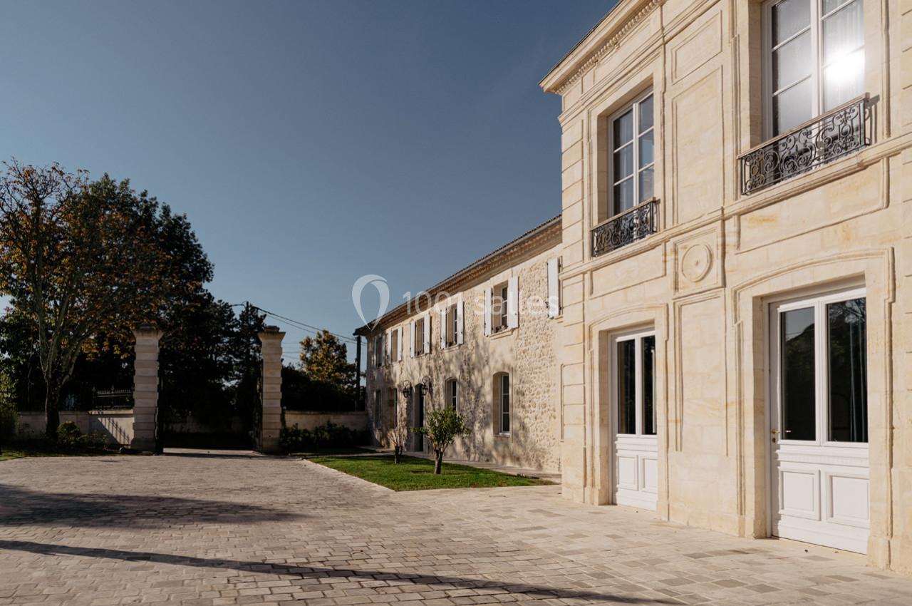 Façade d'une maison en pierre avec des fenêtres blanches, une cour pavée et des arbres sous un ciel dégagé.