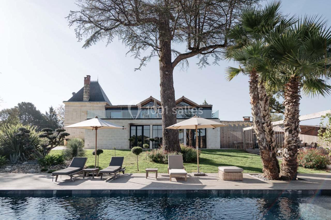 Jardin avec piscine, transats et parasols devant une maison en pierre entourée de végétation et de palmiers.