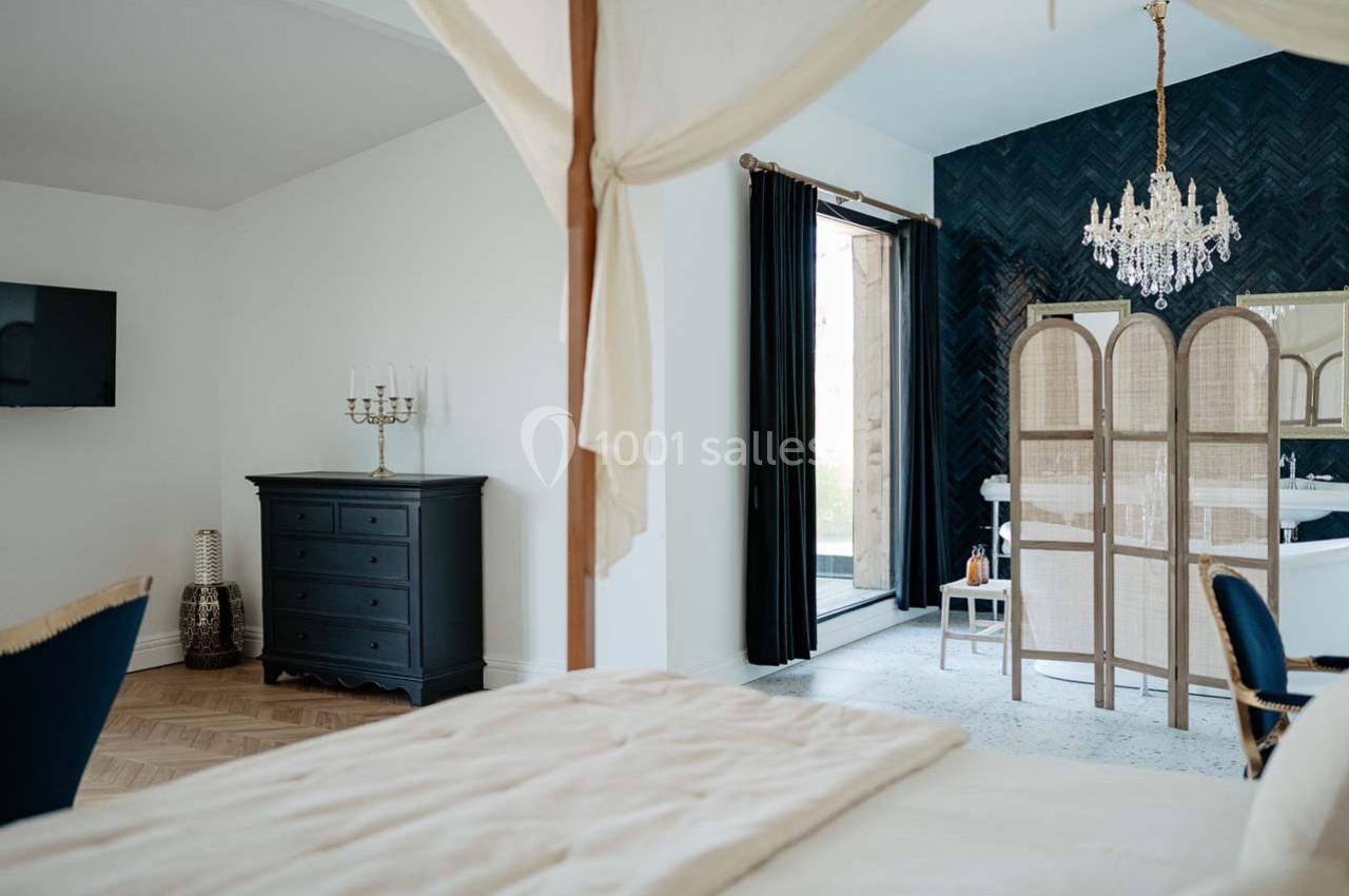 Chambre lumineuse avec lit à baldaquin, commode noire, paravent en rotin, lustre et rideaux sombres.