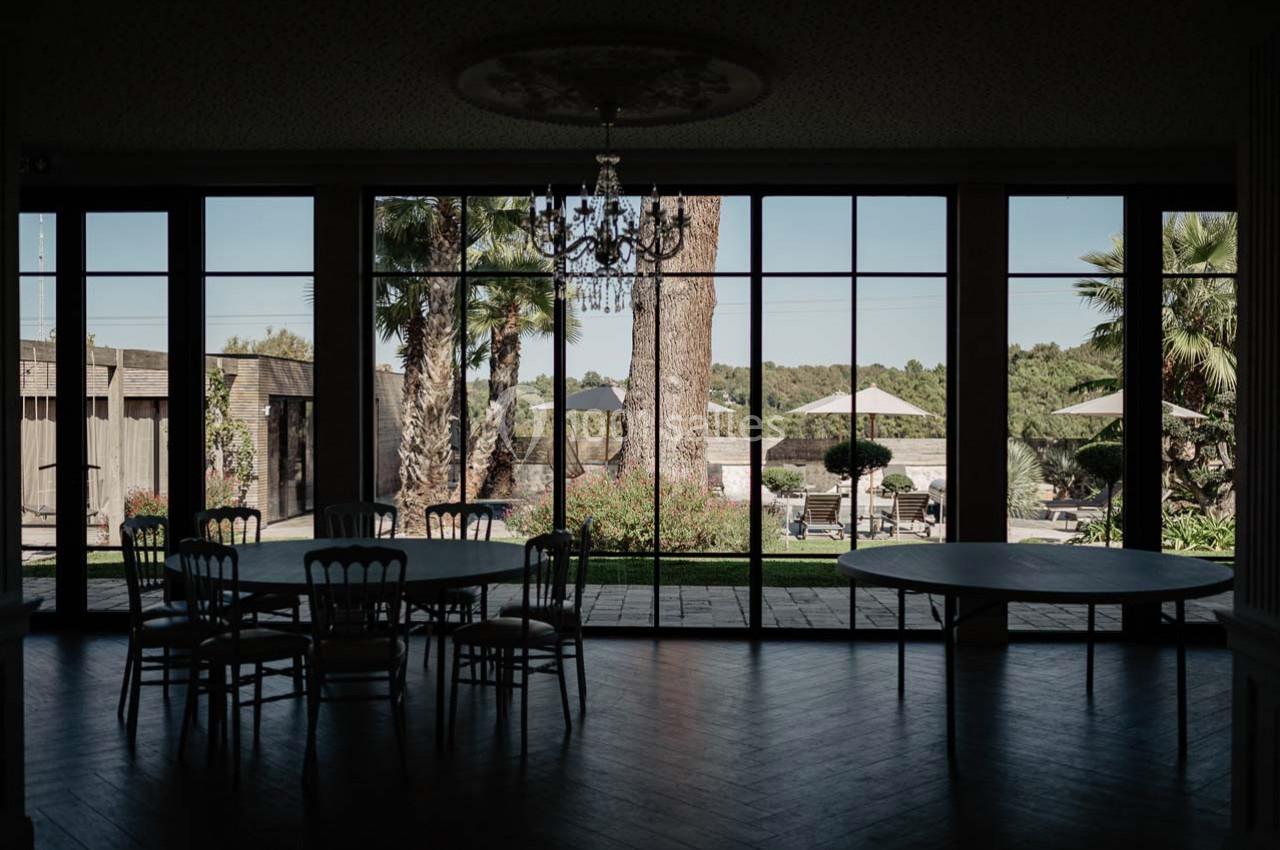 Salle sombre avec des tables et des chaises, donnant sur un jardin ensoleillé avec arbres, parasols et végétation.