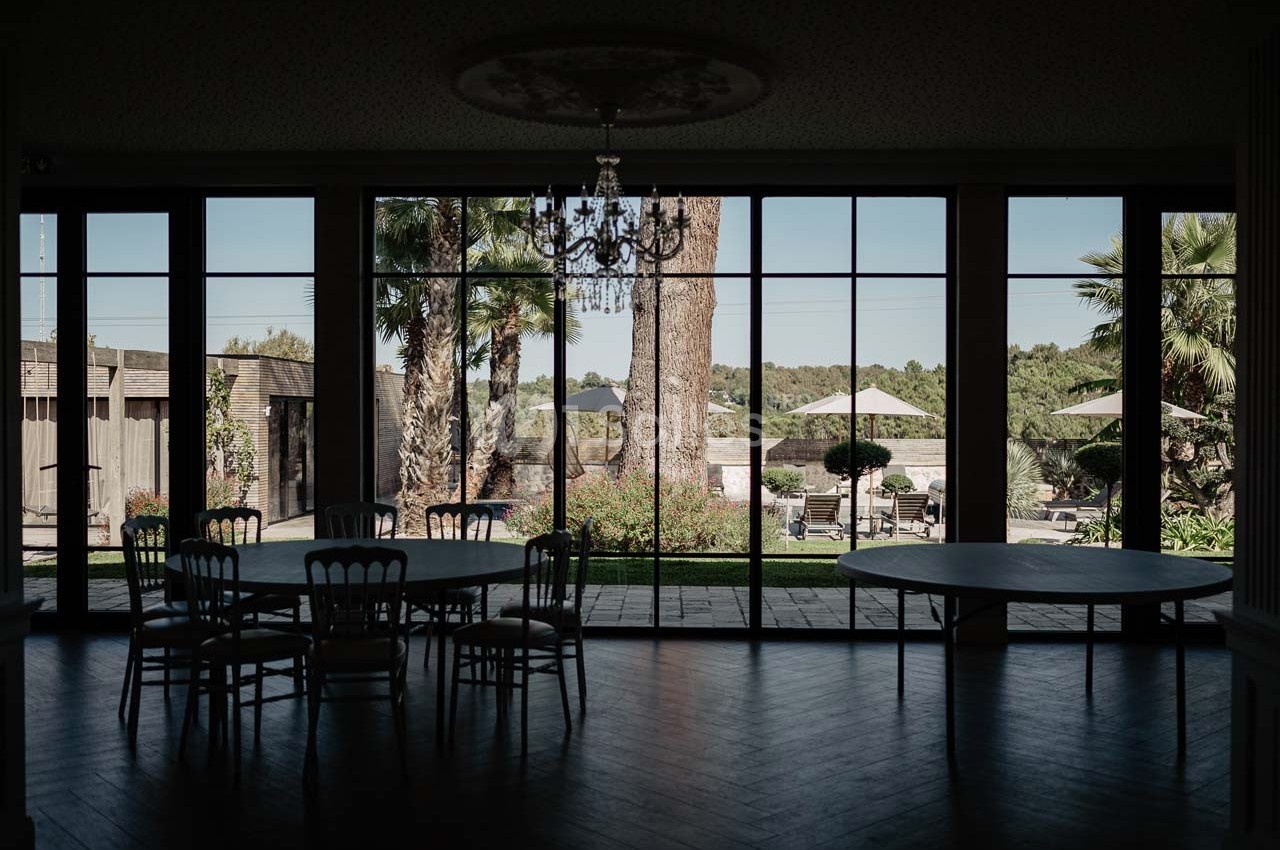 Salle sombre avec des tables et des chaises, donnant sur un jardin ensoleillé avec arbres, parasols et végétation.