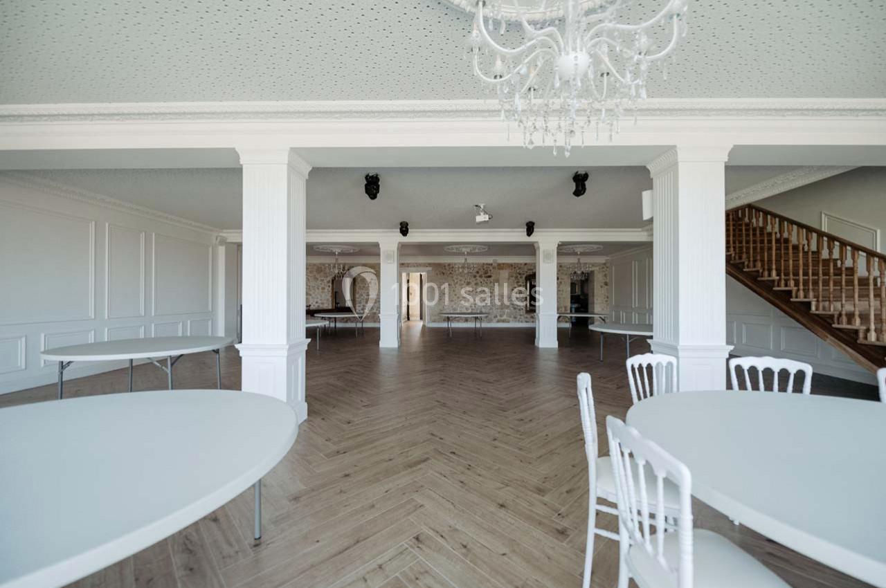 Salle spacieuse avec parquet en chevrons, tables rondes blanches, murs clairs et escalier en bois à droite.