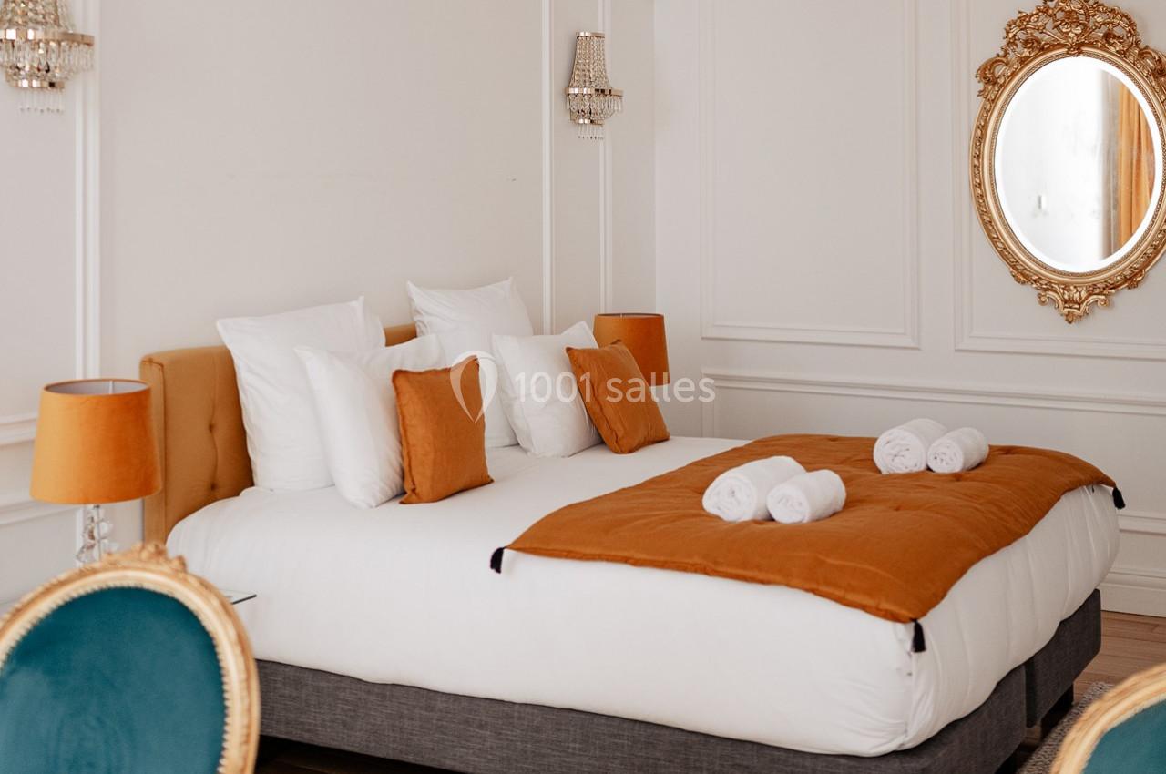 Lit double avec draps blancs, coussins orange, couverture assortie, miroir doré et murs blancs ornés de moulures.