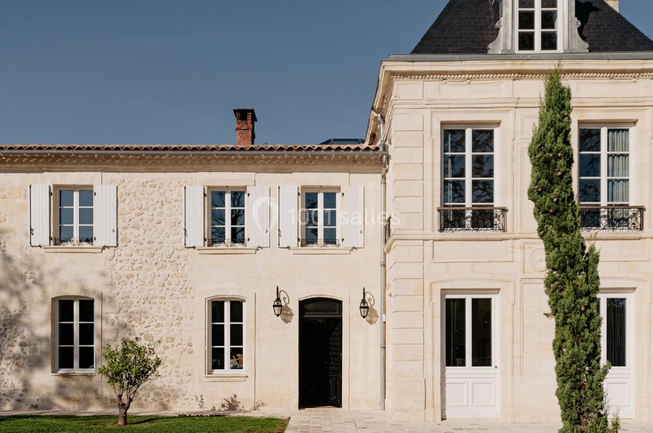 Façade d'une maison en pierre avec volets blancs, toit mansardé et un cyprès dans une cour pavée.