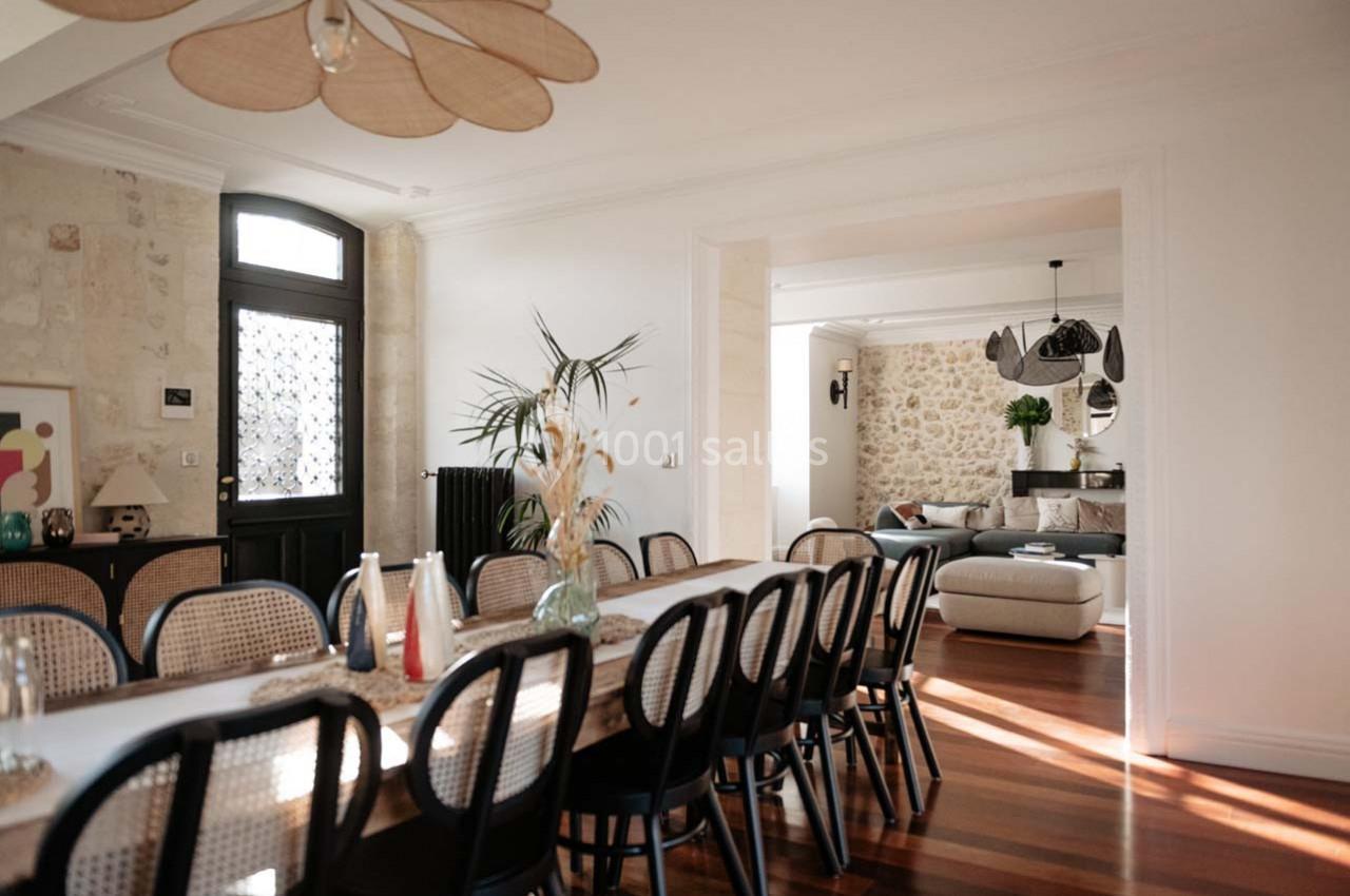 Salle à manger lumineuse avec une grande table en bois, chaises en rotin, et vue sur un salon avec murs en pierre.