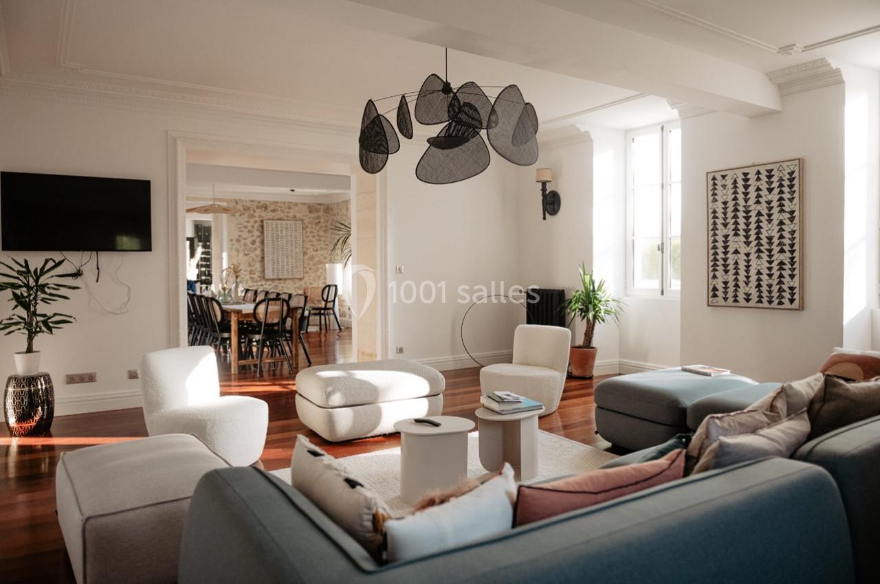 Salon lumineux avec canapé, fauteuils, table basse, suspension design et vue sur une salle à manger en arrière-plan.