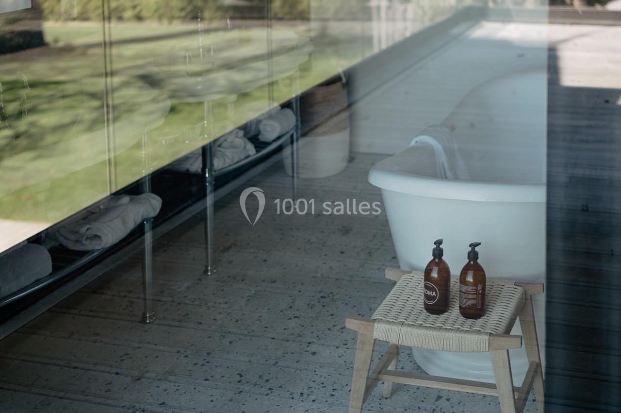 Vue d'une salle de bain avec une baignoire blanche, un tabouret en bois portant deux flacons, et une vitre donnant sur un…