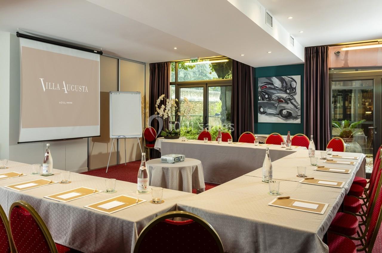 Salle de réunion lumineuse avec tables disposées en U, écran de projection, paperboard et vue sur un jardin.