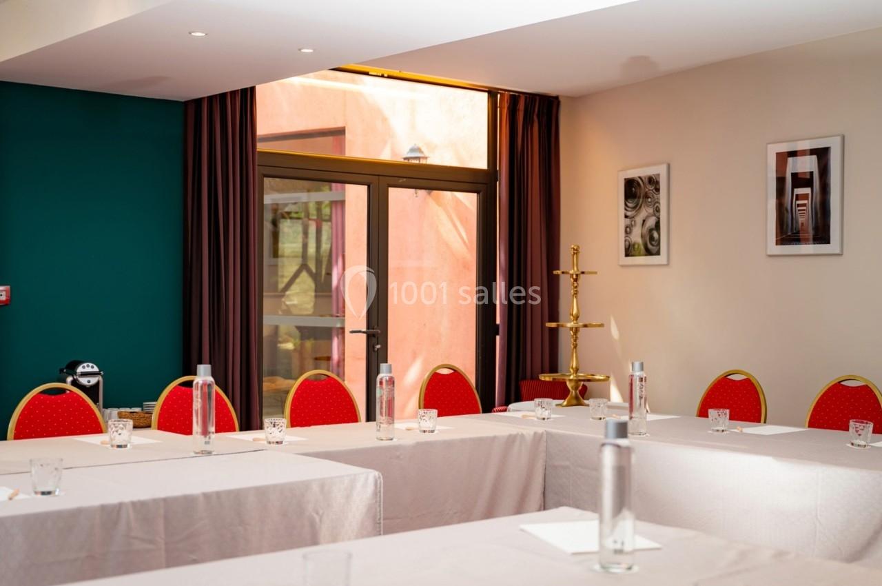 Salle de réunion lumineuse avec tables disposées en U, chaises rouges, bouteilles d'eau et fenêtres donnant sur l'extérieur.