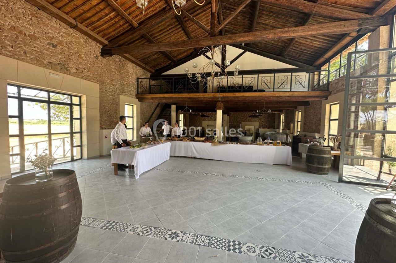 Salle spacieuse avec murs en pierre, plafond en bois, grandes fenêtres et tables dressées pour un buffet.