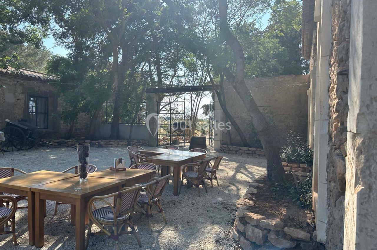 Cour extérieure ensoleillée avec tables en bois, chaises en osier, arbres et vue sur un portail en fer forgé.