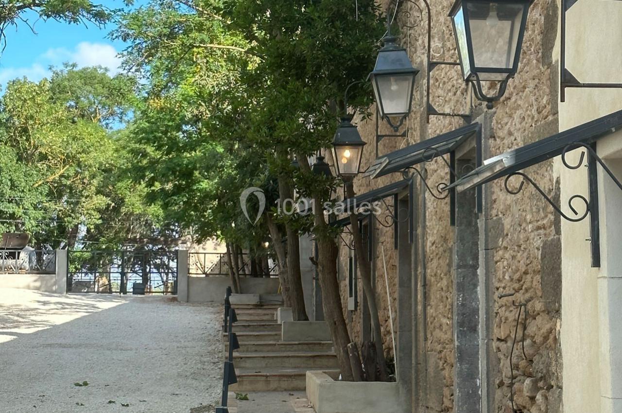 Façade en pierre avec lanternes murales, bordée d'arbres et d'un chemin en gravier menant à une ouverture lumineuse.
