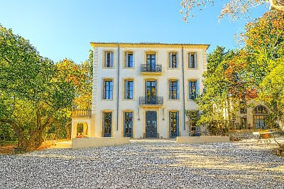 Un couple en tenue de mariage marche main dans la main devant un bâtiment en pierre et des arbres.
