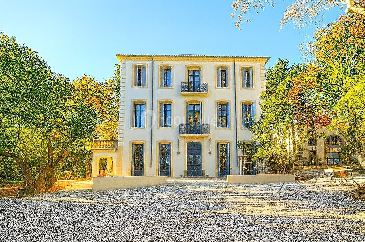 Grande maison blanche à trois étages entourée d'arbres, avec une cour en gravier sous un ciel dégagé.