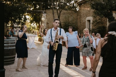 Un couple en tenue de mariage marche main dans la main devant un bâtiment en pierre et des arbres.