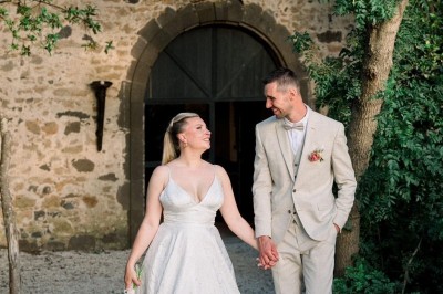 Un couple en tenue de mariage marche main dans la main devant un bâtiment en pierre et des arbres.