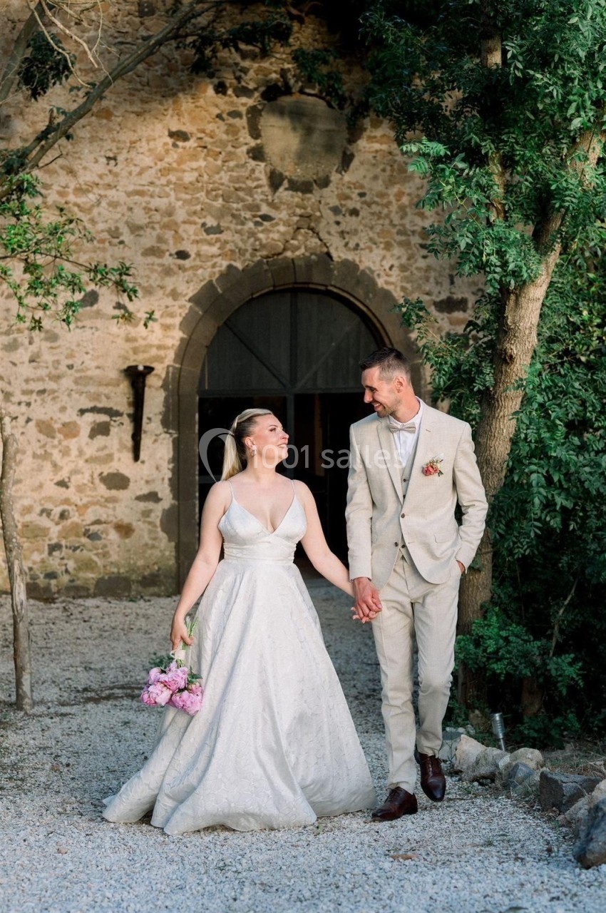 Un couple en tenue de mariage marche main dans la main devant un bâtiment en pierre et des arbres.