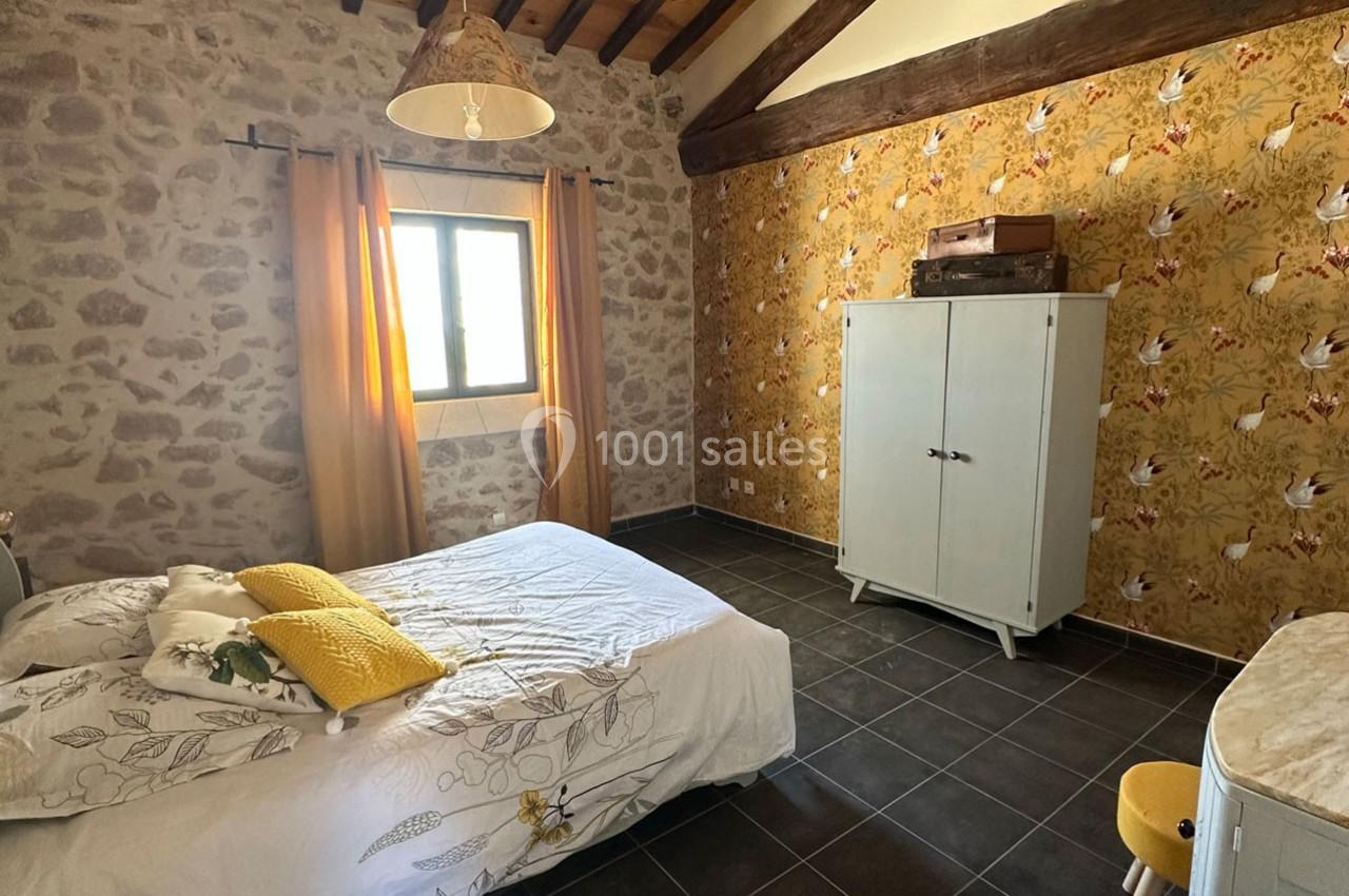 Chambre avec murs en pierre et papier peint à motifs, lit double, armoire blanche et fenêtre laissant entrer la lumière.