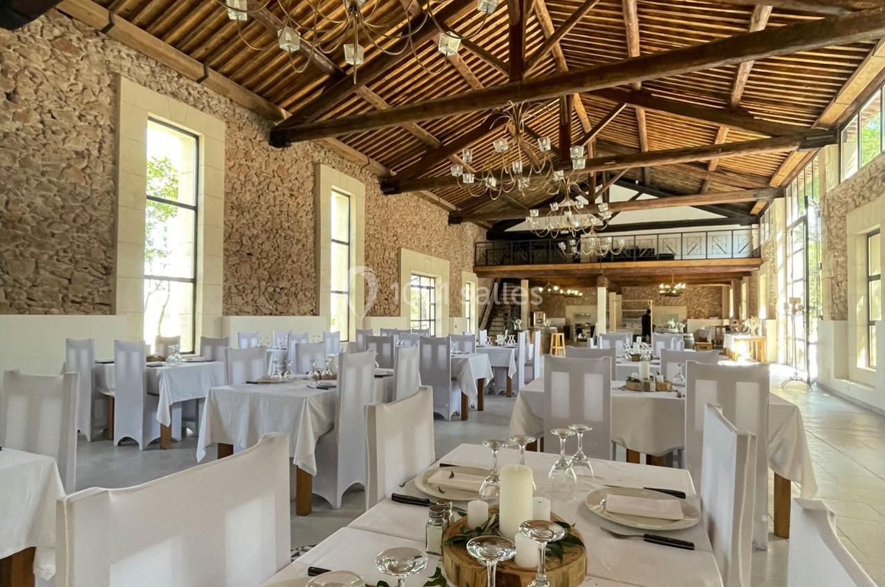 Salle de réception lumineuse avec tables dressées, chaises blanches et poutres apparentes sous un toit en bois.