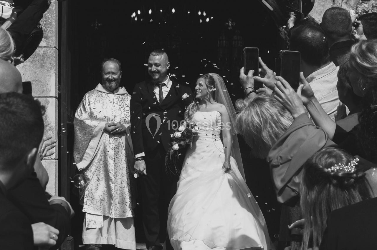 Un couple de mariés sort d'une église, entouré d'invités tenant des ballons et prenant des photos.