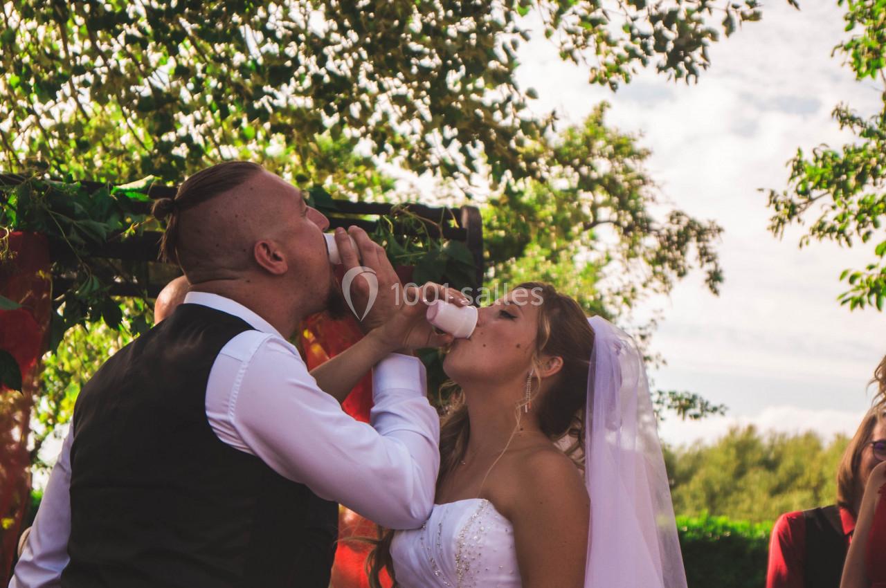 Un couple en tenue de mariage boit ensemble dans une corne lors d'une cérémonie en extérieur, entouré d'invités.
