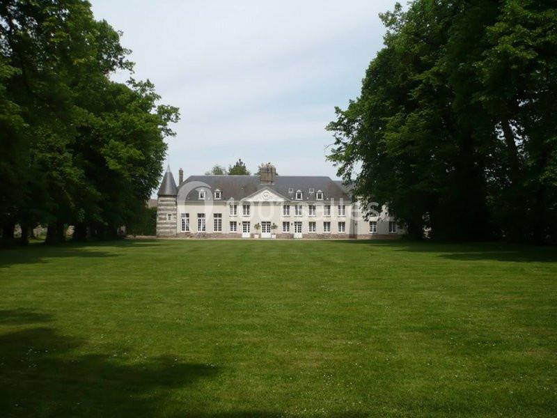 Façade d'un manoir blanc entouré de pelouses et d'arbres sous un ciel légèrement nuageux.