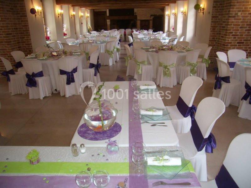 Salle de réception décorée pour un événement, avec des tables dressées et des chaises ornées de nœuds violets.