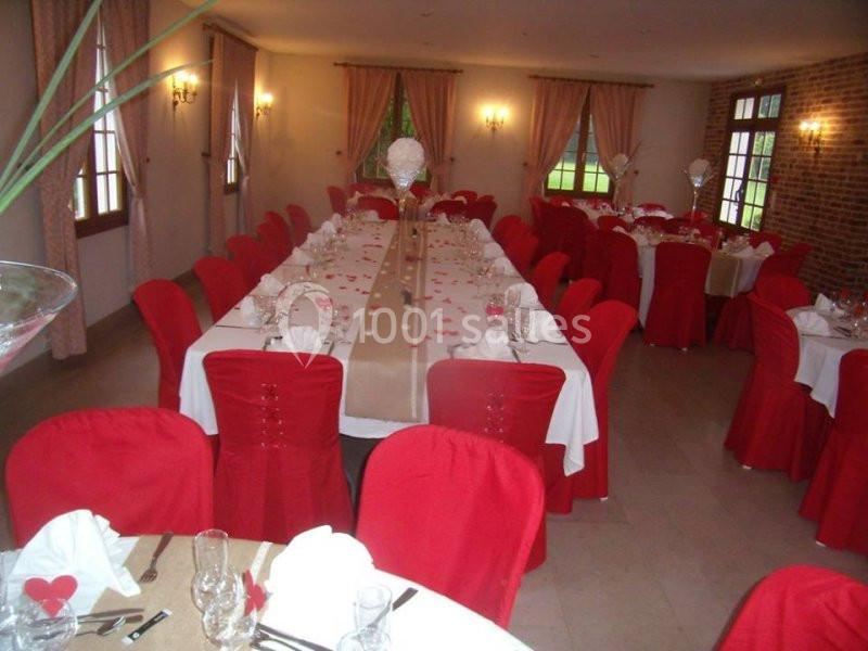Salle de réception avec deux longues tables dressées, nappes blanches, chaises rouges et décoration florale centrale.