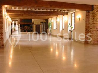 Salle spacieuse avec sol en pierre, poutres apparentes au plafond, murs en briques et grandes fenêtres.