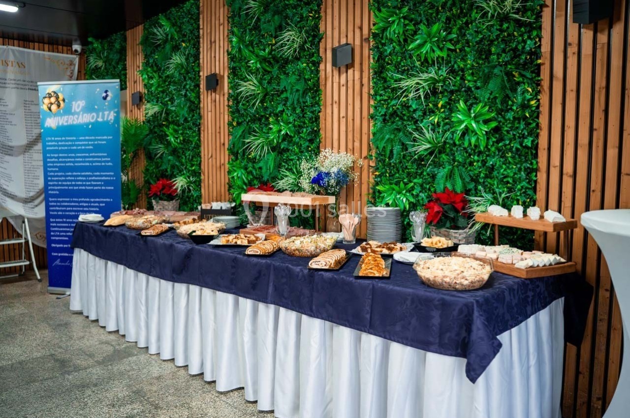 Buffet varié avec pâtisseries, plats salés et desserts, disposé sur une table drapée, devant un mur végétalisé.