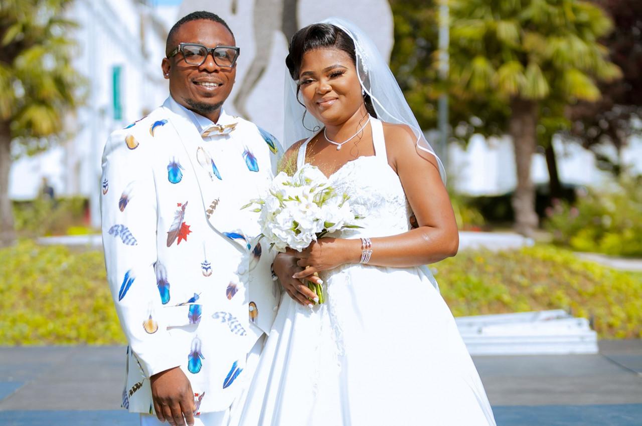 Un couple souriant en tenue de mariage pose à l'extérieur devant un décor verdoyant et lumineux.