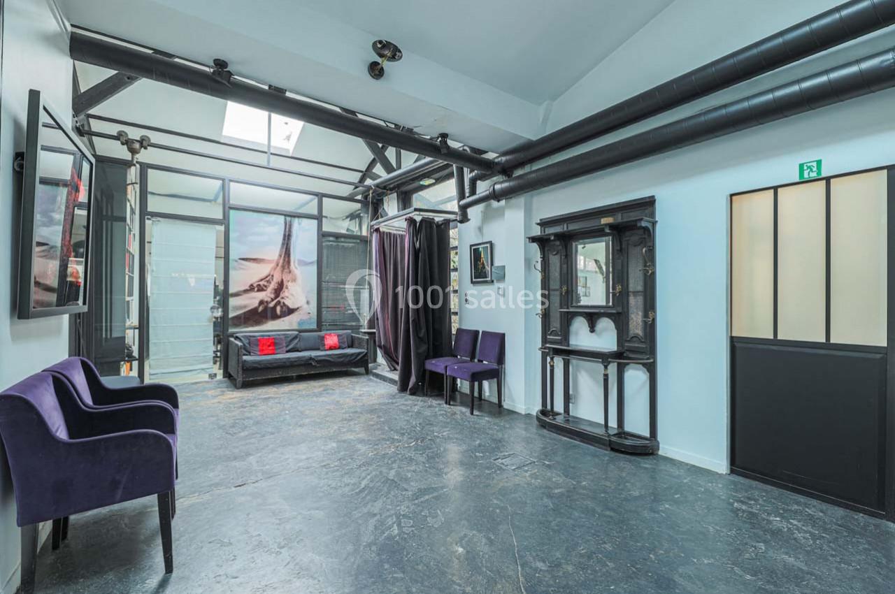 Espace intérieur lumineux avec murs blancs, sol en béton, mobilier violet et noir, et grandes baies vitrées.