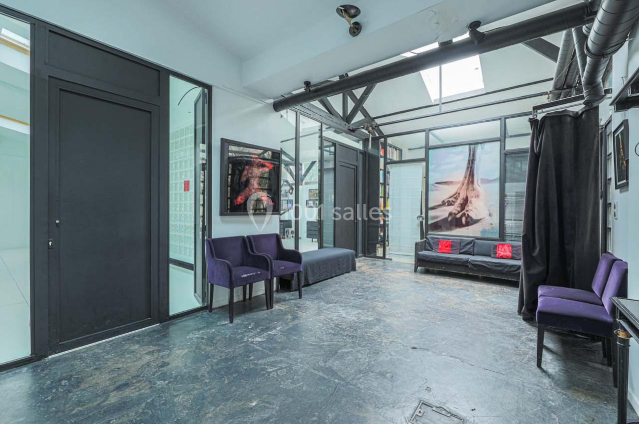 Salle lumineuse avec sol en béton, fauteuils violets, grandes baies vitrées et décoration murale artistique.