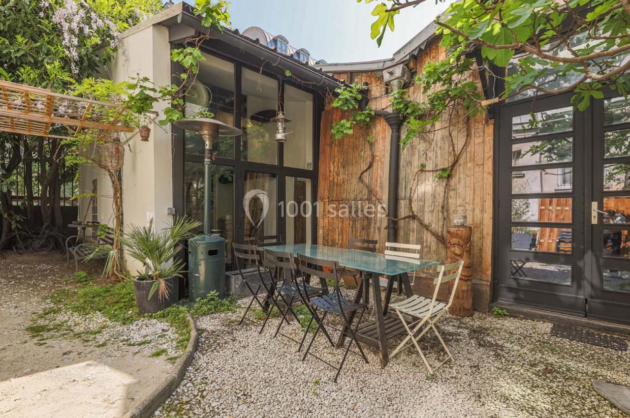 Terrasse extérieure avec table en verre, chaises et végétation grimpante, entourée de murs en bois et baies vitrées.