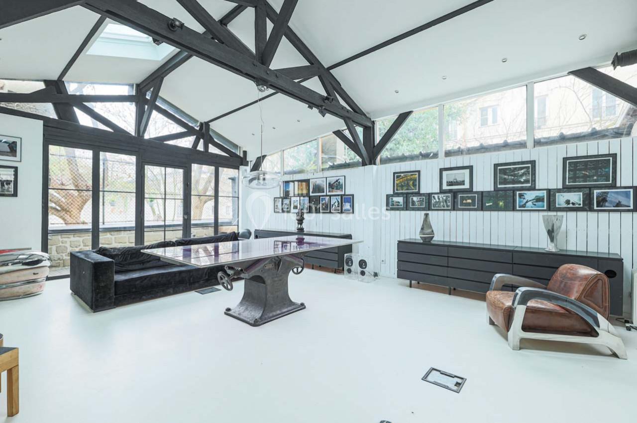 Salle lumineuse avec charpente apparente, table de ping-pong, canapés, fauteuils et murs décorés de cadres photo.