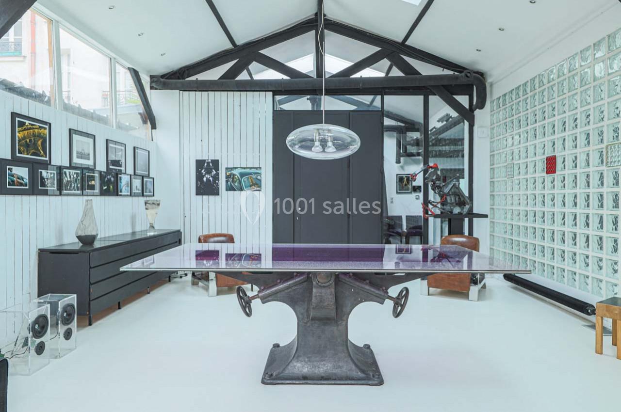 Salle lumineuse avec une grande table au pied métallique, entourée de chaises, décorée de photos et d'objets modernes.