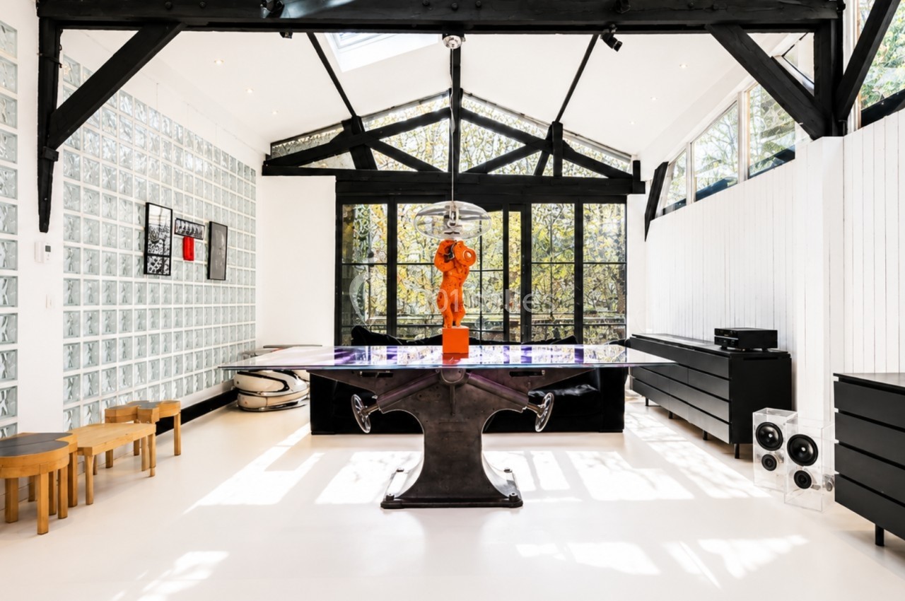 Salle lumineuse avec verrière, table design noire, sculpture orange centrale, murs blancs et mobilier moderne.