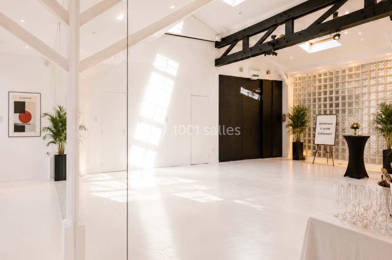 Salle lumineuse avec sol blanc, poutres noires apparentes, plantes décoratives et table dressée pour un événement.
