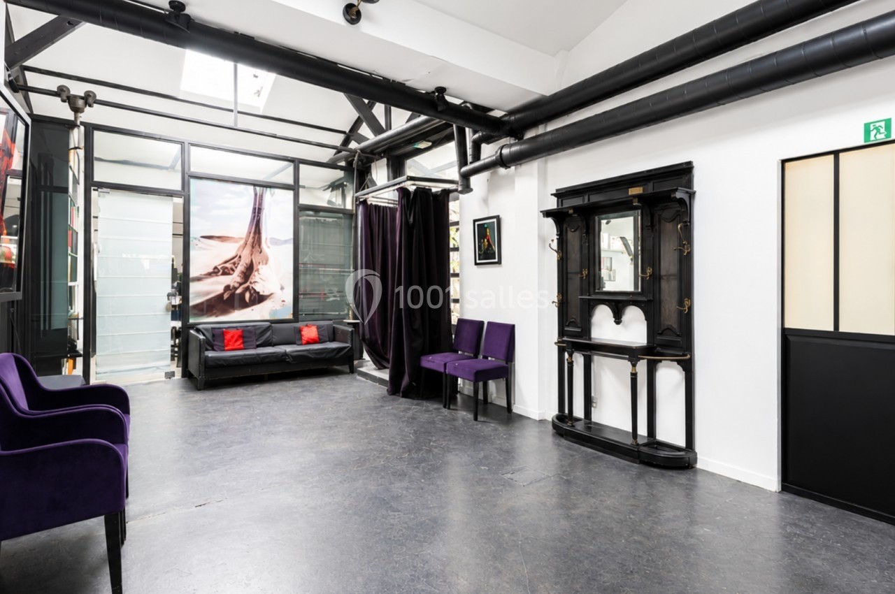 Salle lumineuse avec sol en béton, mobilier violet et noir, et grande baie vitrée donnant sur une autre pièce.