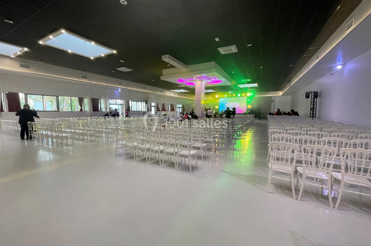Salle de réception spacieuse avec des rangées de chaises blanches alignées face à une scène éclairée par des lumières…