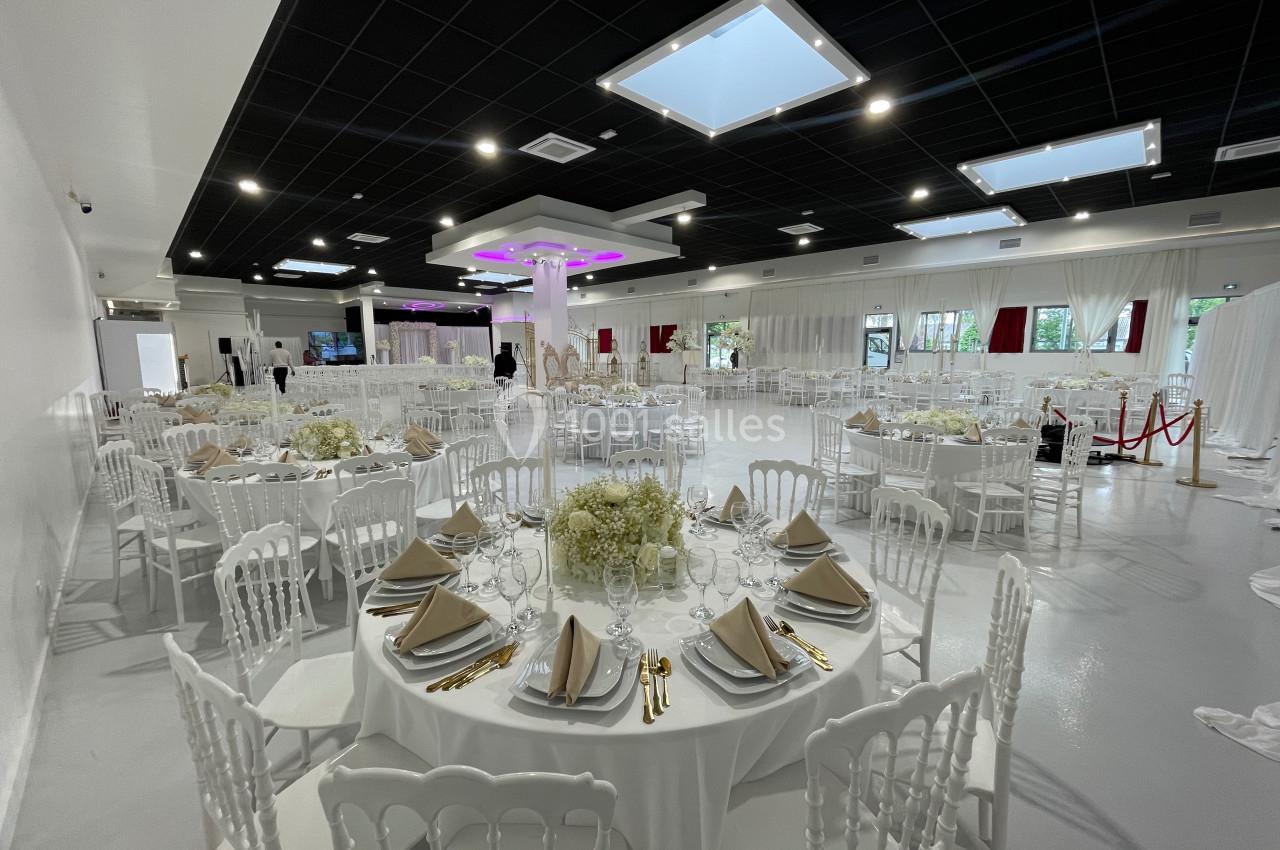 Salle de réception élégante avec tables rondes dressées, nappes blanches, chaises blanches et décorations florales.