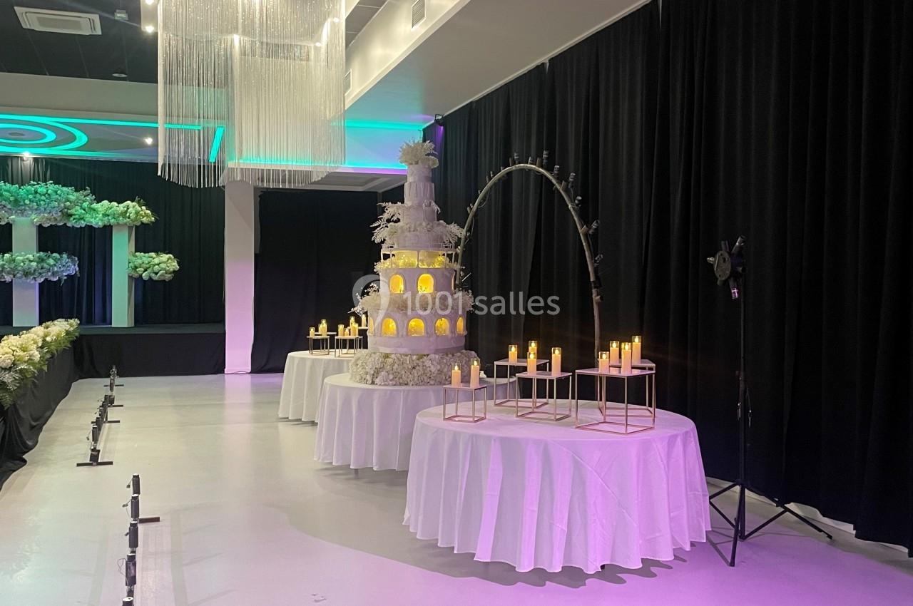 Salle de réception décorée avec un grand gâteau de mariage éclairé, des bougies et des rideaux noirs en arrière-plan.