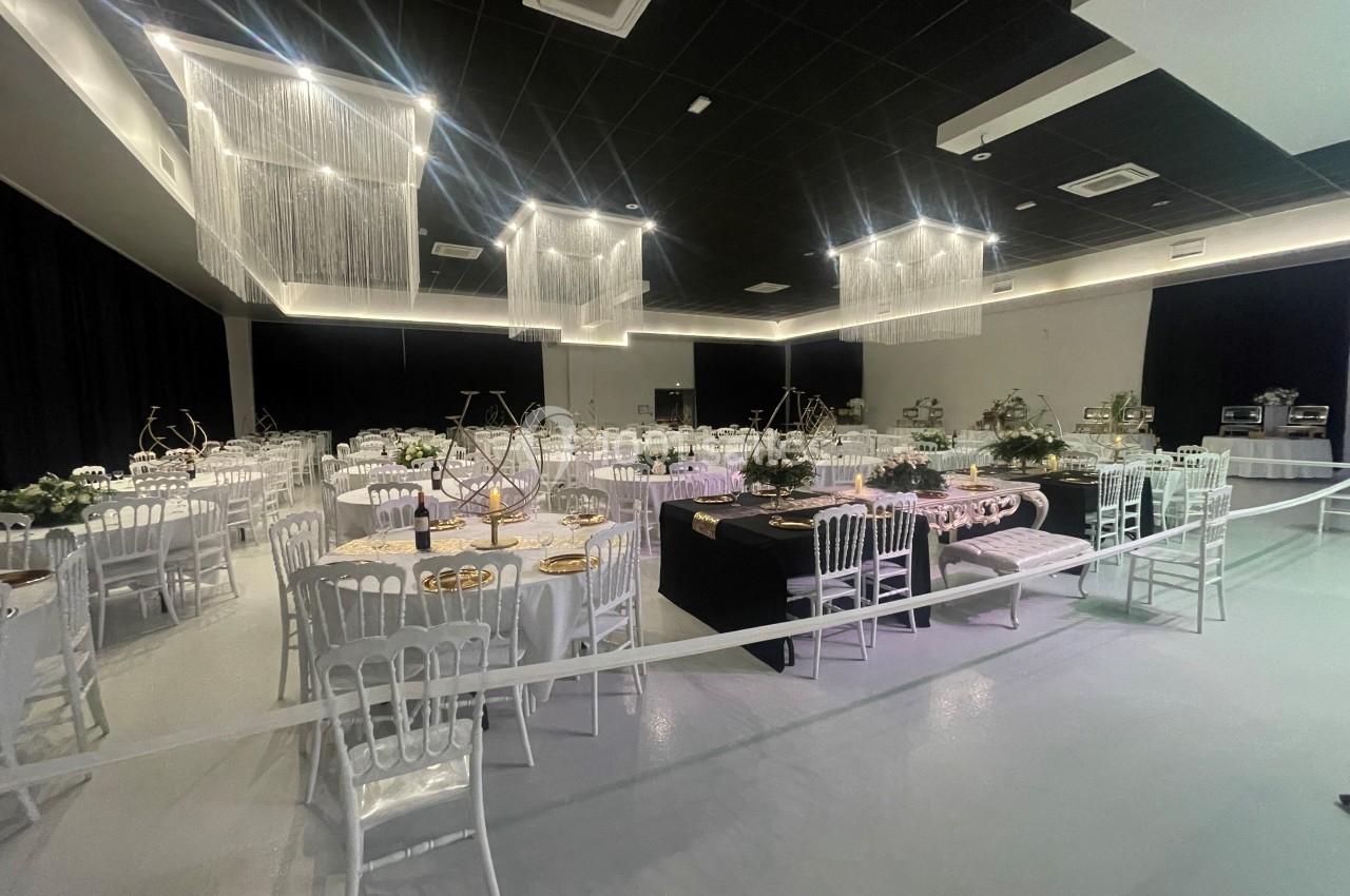 Salle de réception décorée avec des tables rondes, nappes blanches, chaises transparentes et éclairage suspendu.