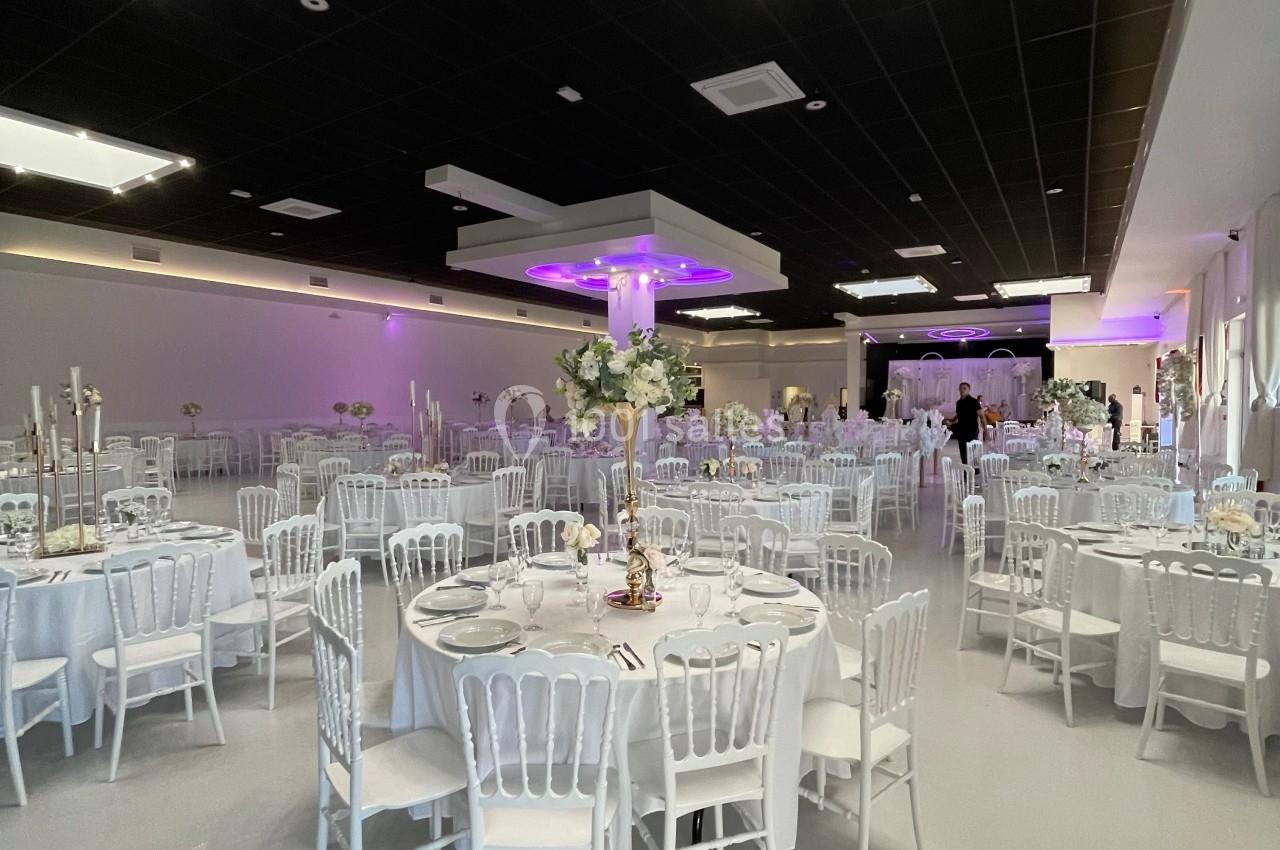 Salle de réception décorée avec des tables rondes dressées, chaises blanches et éclairage tamisé violet.
