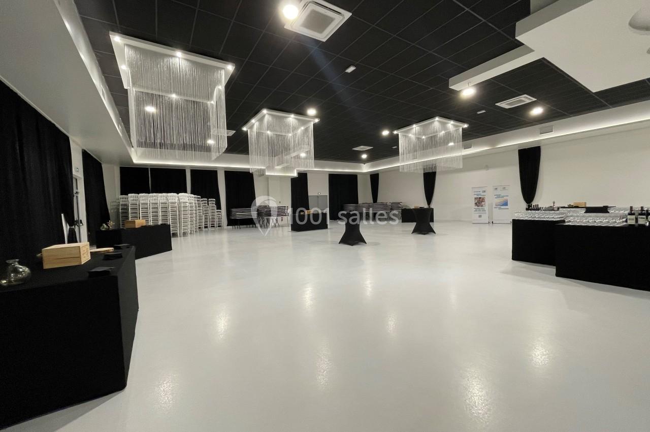 Salle spacieuse avec sol clair, tables hautes noires, chaises empilées et éclairage moderne suspendu.