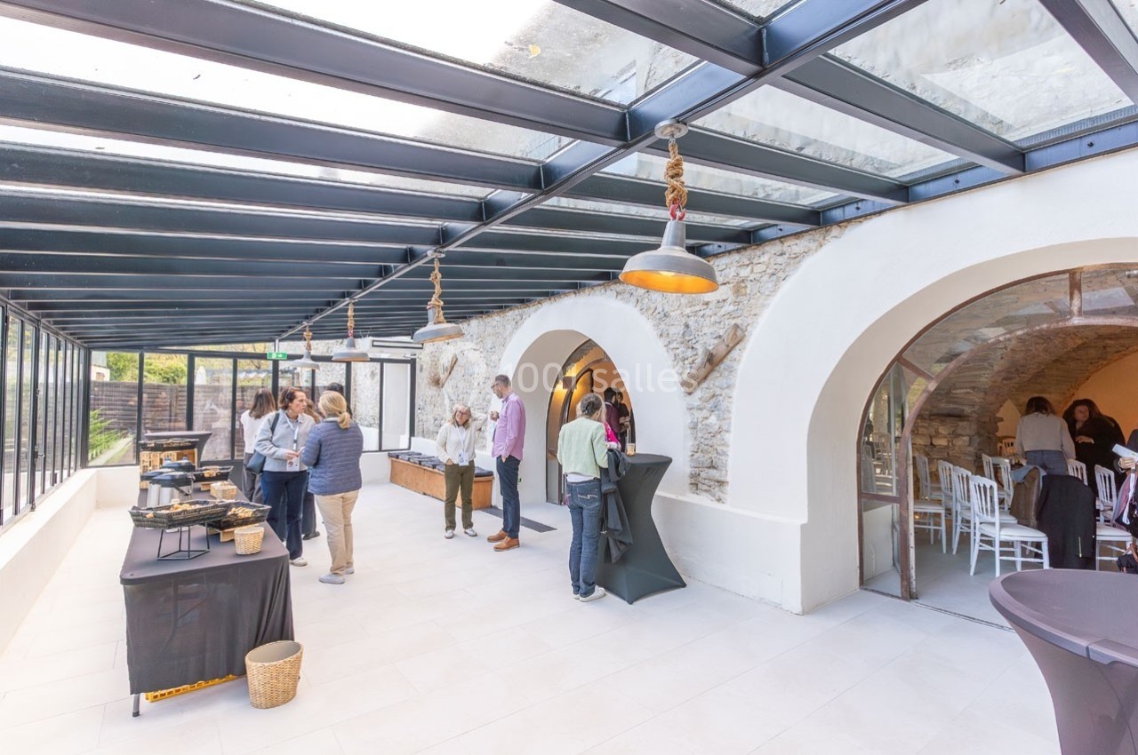 Salle lumineuse avec verrière, murs en pierre, tables de buffet et personnes discutant ou se servant.