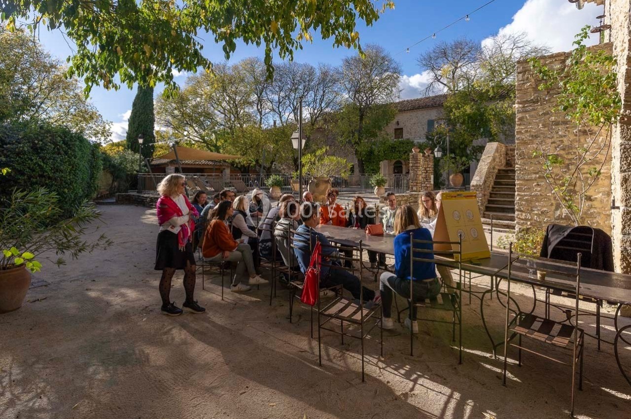 Un groupe de personnes assises autour d'une table en extérieur dans une cour ensoleillée entourée de bâtiments en pierre.