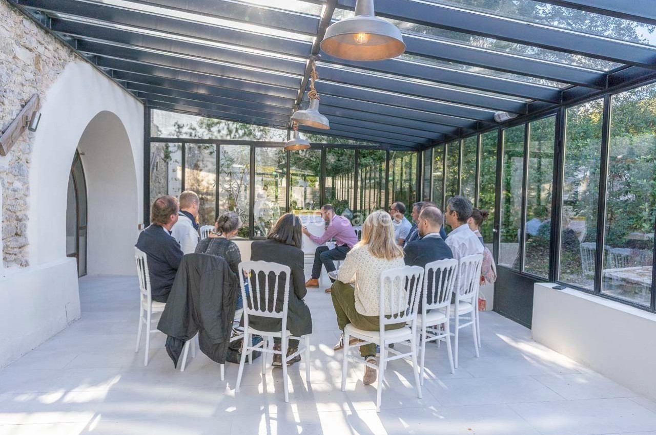 Un groupe de personnes assises en cercle dans une véranda lumineuse avec des murs en verre et vue sur un jardin.